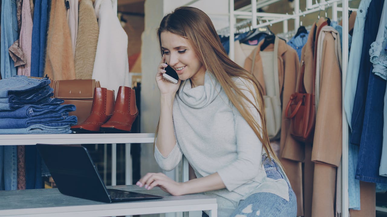 Smiling woman in a fashion boutique using a laptop and phone.